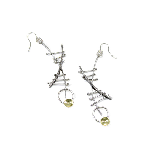 A pair of unique earrings, featuring two textured and arching bars, with one shorter and thicker than the other, crossing over each other to create an X shape. Three bars on the top and bottom halves of the X respectively run between the gap of the arches. Each bar's end pierces through the frame of the X, secured by a silver ball. At the bottom of the longest bar, a silver ring hangs, adorned with a gold cup sitting on its edge. Custom-made ear hooks are attached to the top of the X