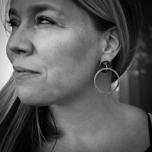 Monochrome side profile of a woman's face wearing a modern handmade silver earring. The earring features a textured, brutalist disc with a smaller ring in the center and a large hoop hanging from the ring, reminiscent of a door knocker